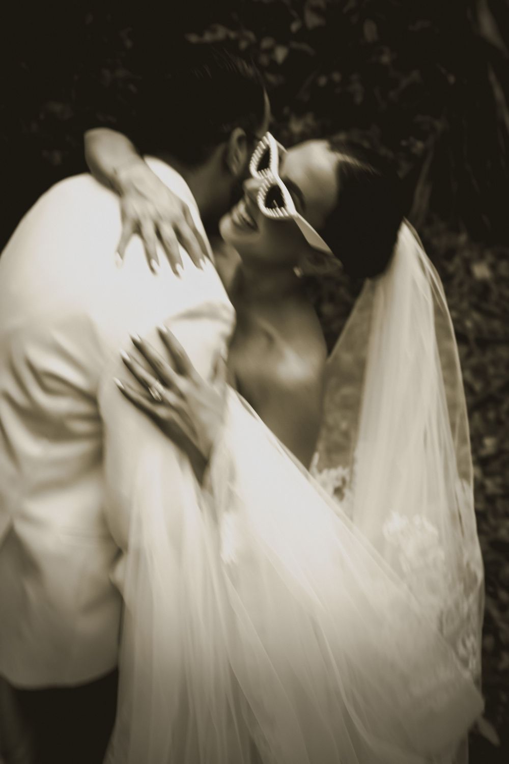 Sepia-toned wedding photo of bride in veil and groom in white suit sharing romantic moment outdoors.