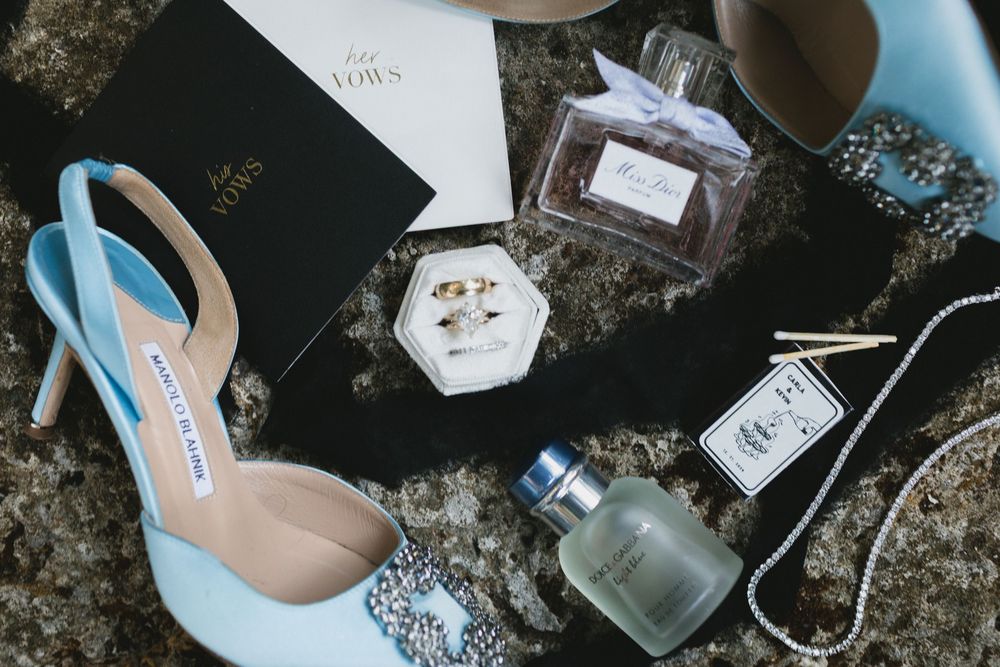 Flat lay of bridal accessories including blue Manolo Blahnik heels, wedding rings, perfume bottles, and pearls.