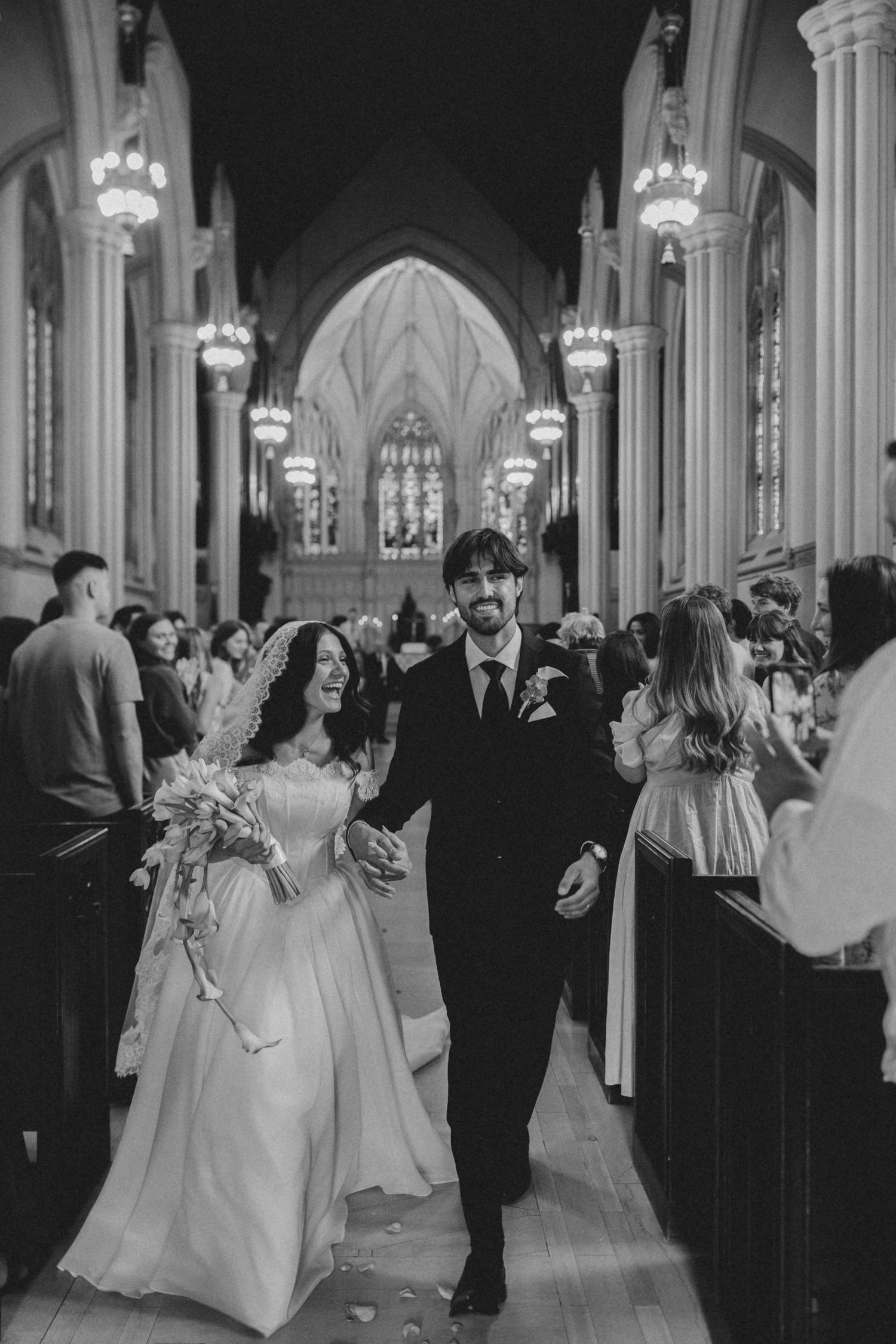 Black and white photo of a bride and groom walking down the aisle of a grand cathedral after their wedding ceremony.