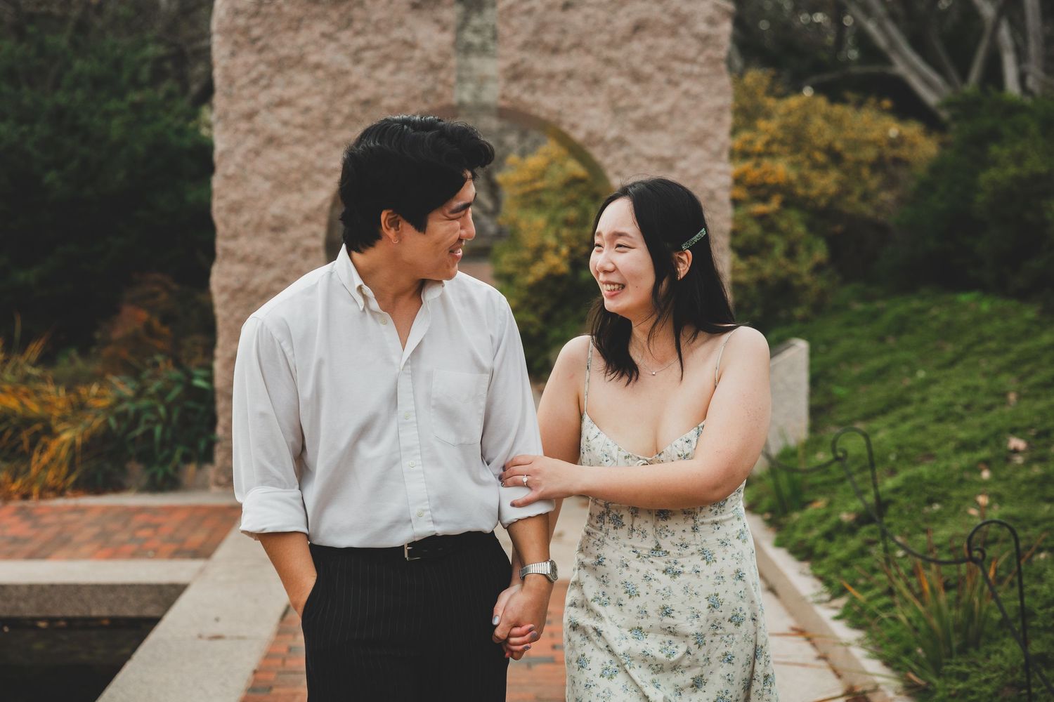 A smiling Asian couple holds hands and gazes at each other in a lush garden setting, man in white shirt, woman in floral dress.