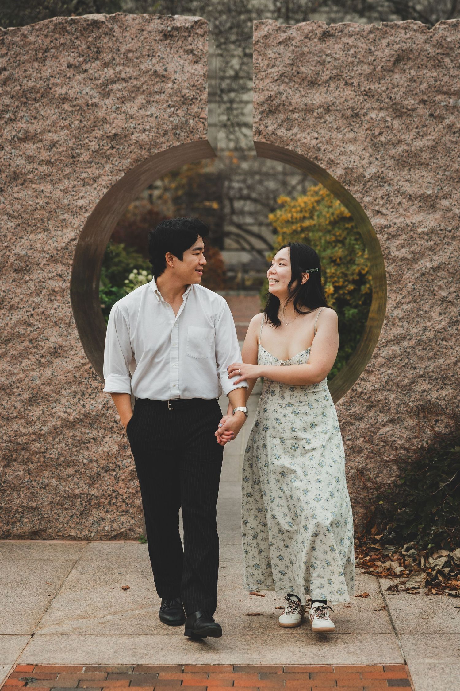 Couple holding hands walking past a circular stone garden gate, smiling at each other in an outdoor setting.