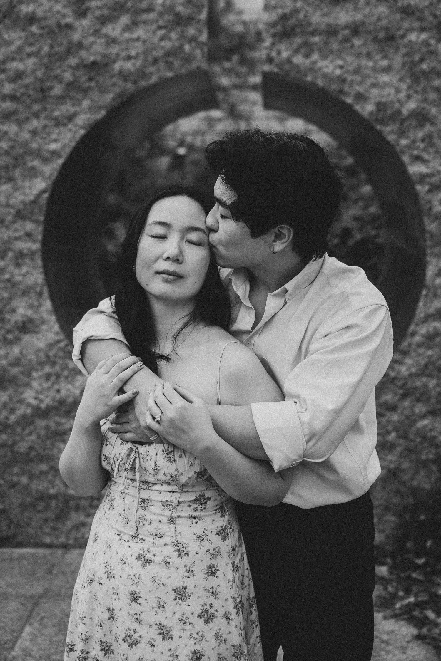 Black and white photo of a couple embracing, man kissing woman on cheek near circular stone arch backdrop.