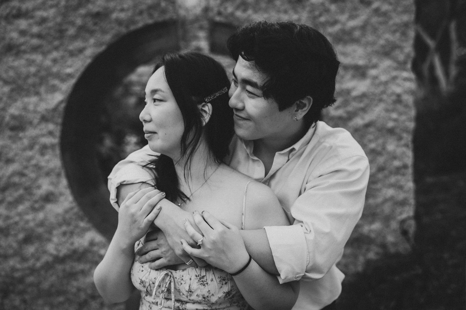 Black and white photo of a couple embracing tenderly outdoors near a stone wall with a circular window.