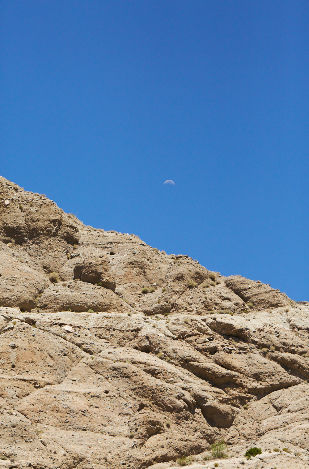 A rugged desert rock formation under a clear blue sky with a half moon visible above the sandy limestone cliffs.