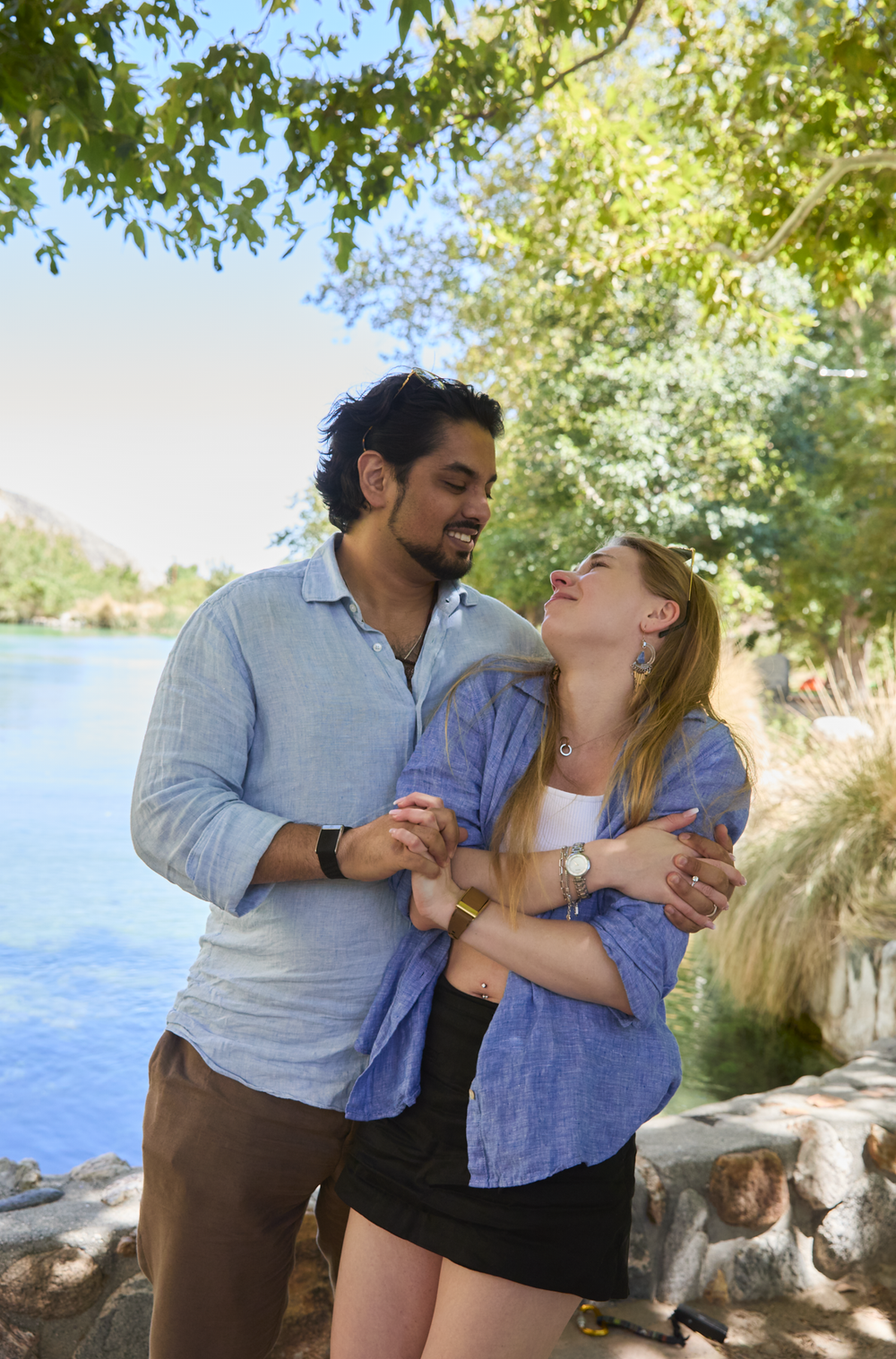 A couple embraces and smiles by a scenic lake surrounded by lush green trees on a sunny day.