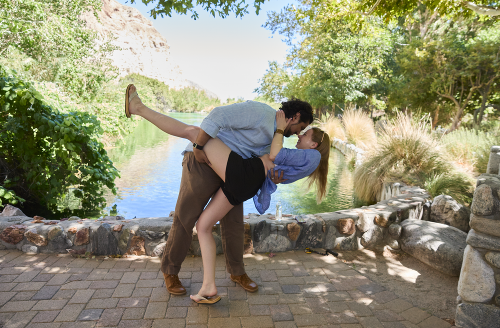 Couple sharing a romantic dip kiss outdoors near a scenic pond surrounded by lush green trees.