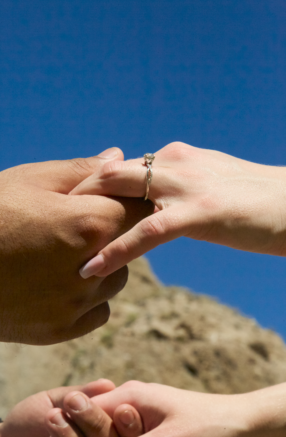 Two hands touching with a diamond engagement ring on finger against blue sky and rocky background.