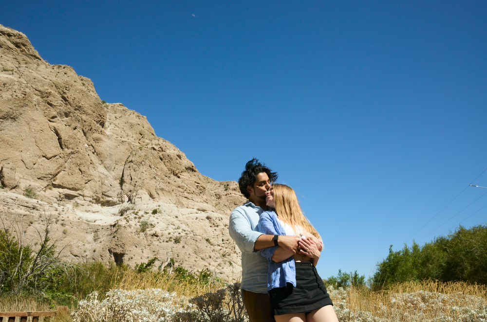 Couple embracing romantically outdoors near sandy rock formations under a clear blue sky.