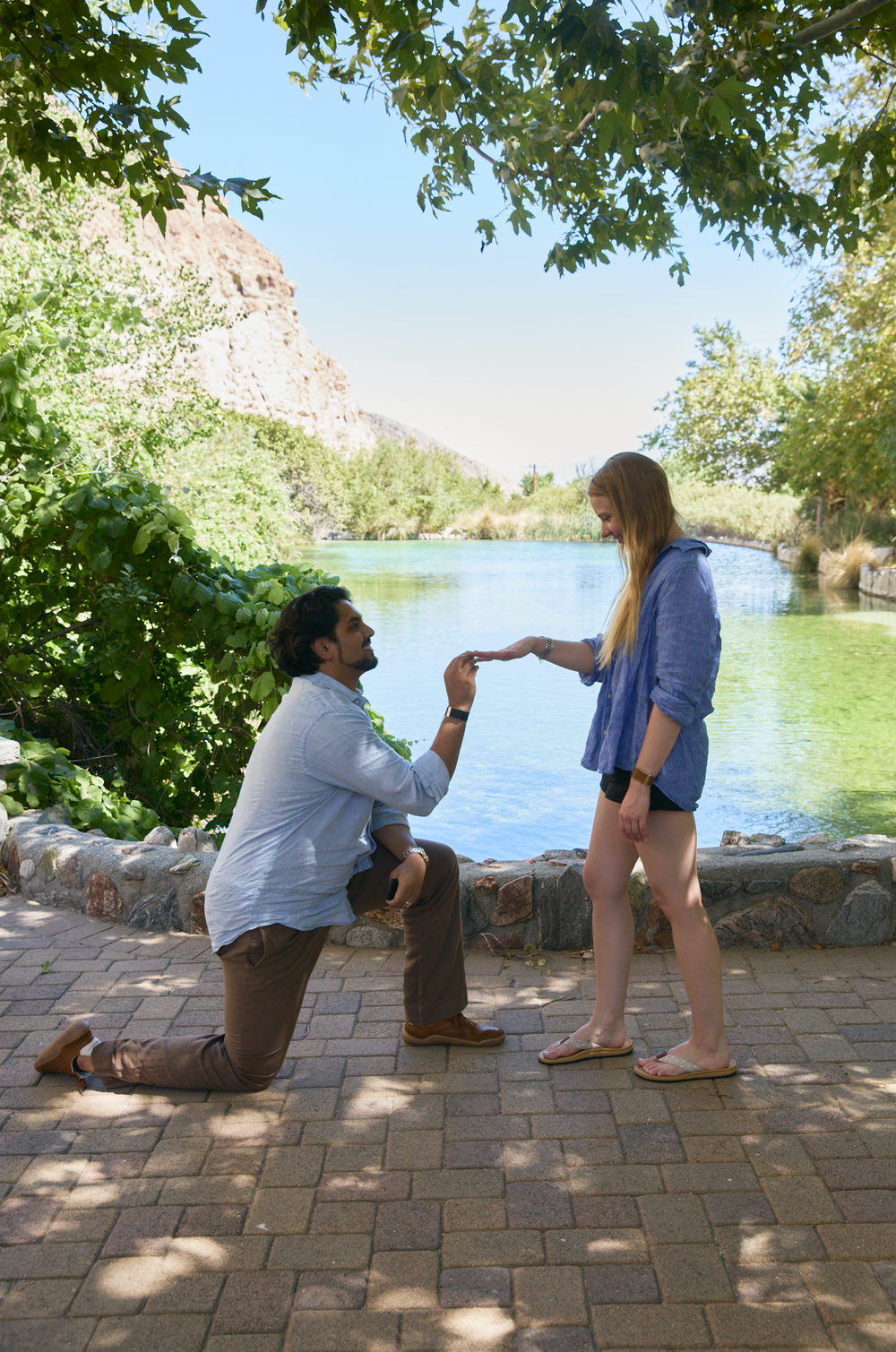 Man kneeling on one knee proposing to woman by scenic turquoise lake surrounded by lush green trees.