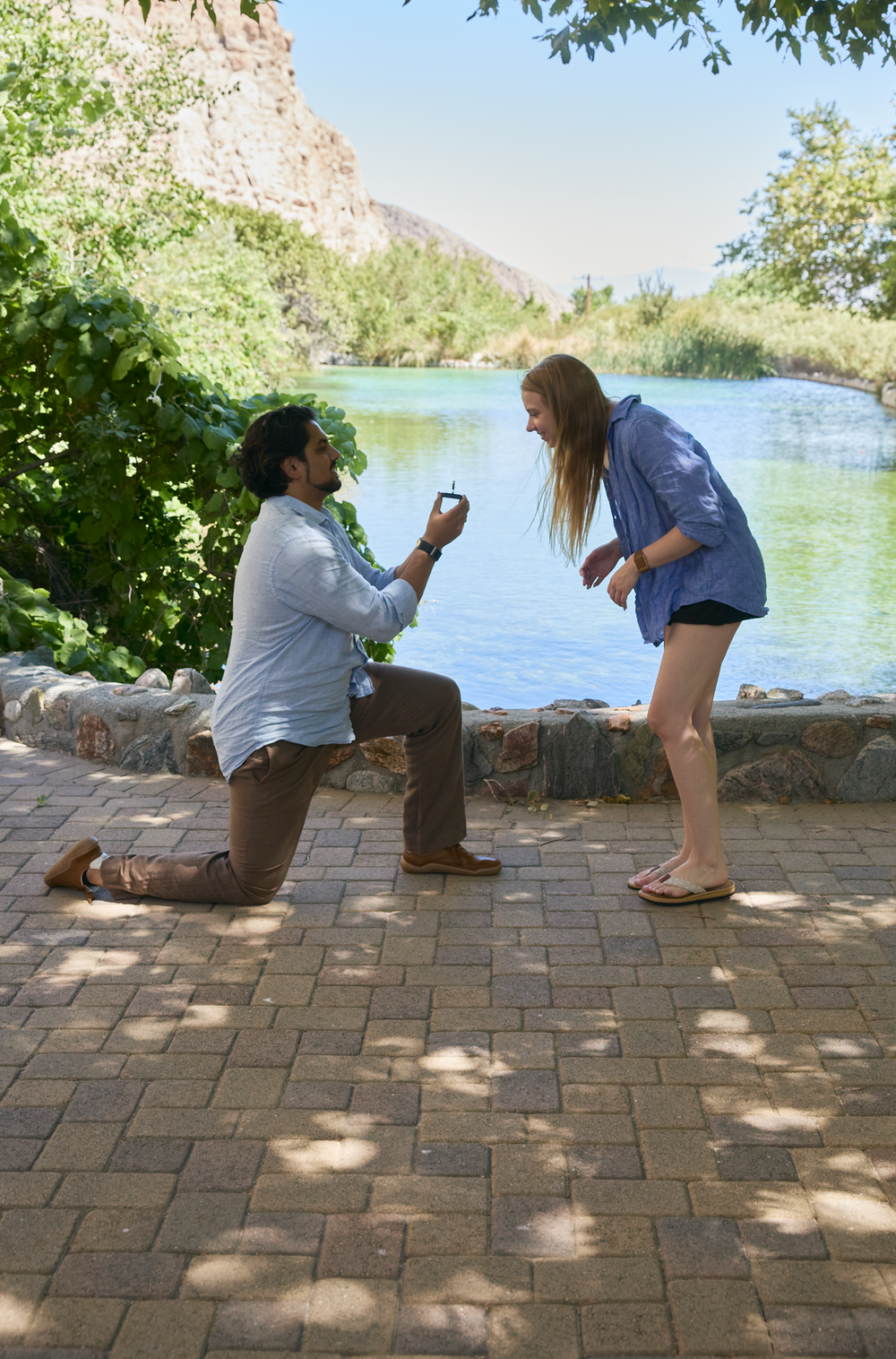 Man kneeling on one knee proposing to woman with ring box by scenic river and green trees.