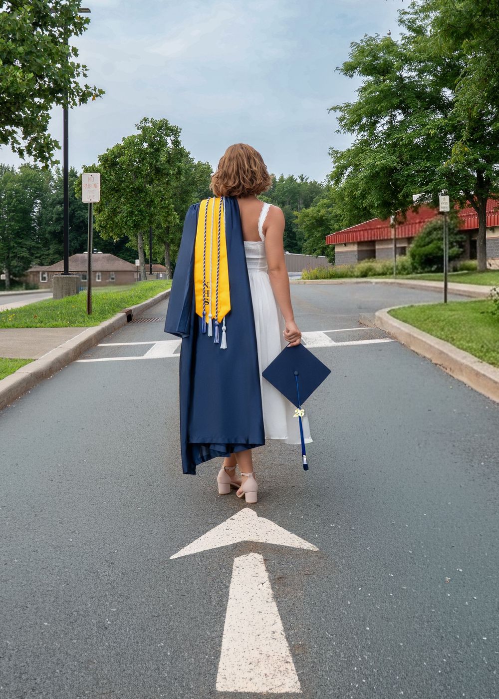 A graduate walks away holding a navy cap and gown with yellow honor cords on a road marked with a forward arrow.