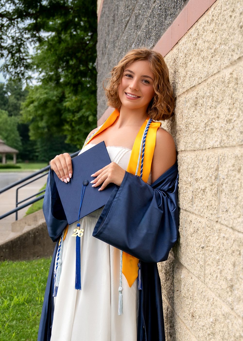 Smiling graduate in navy cap and gown with gold sash holds diploma outdoors near stone building.