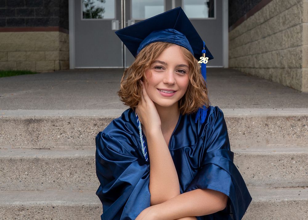 Smiling graduate in blue cap and gown sitting on stone steps for a senior portrait photo.