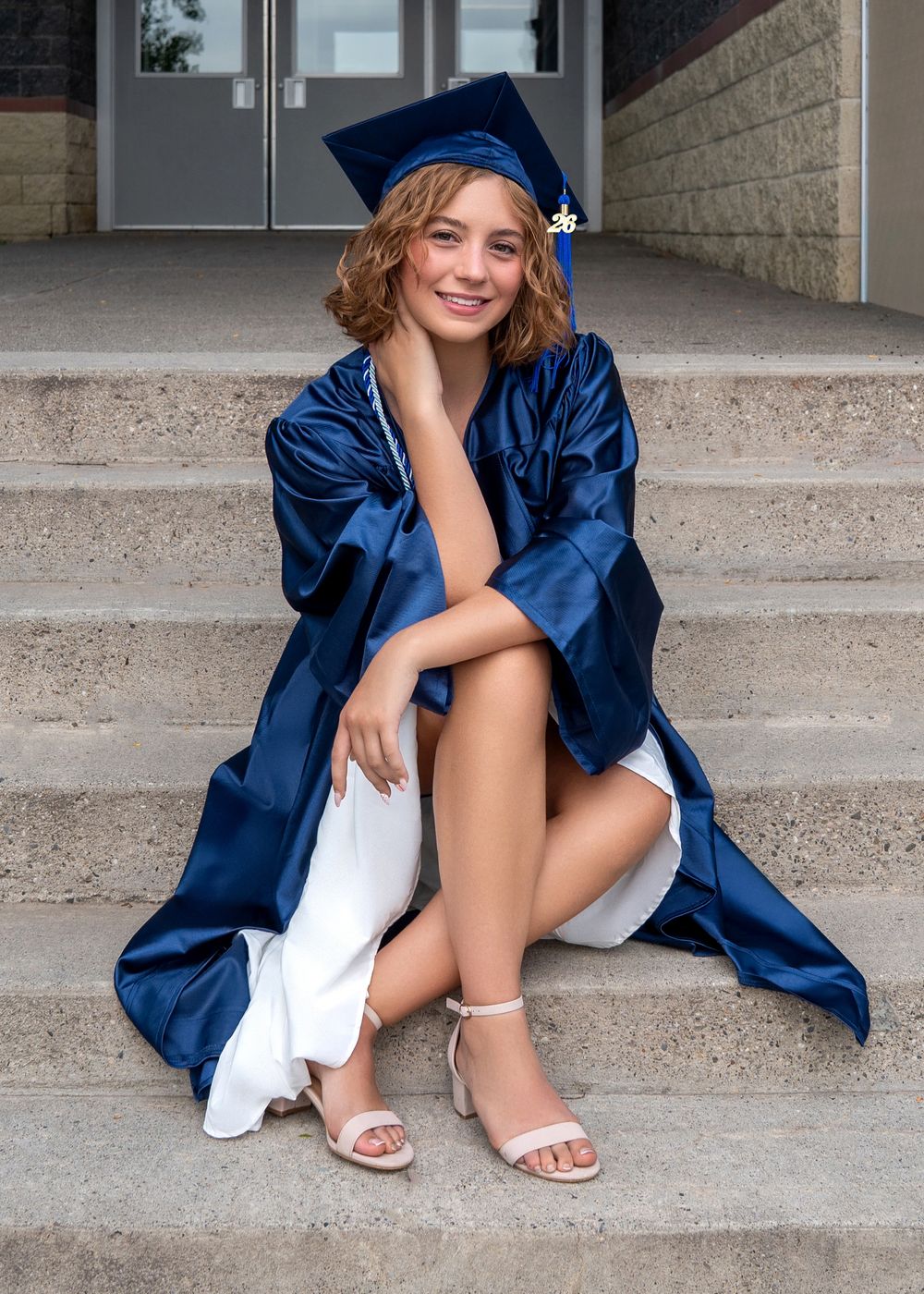 Smiling graduate in blue cap and gown sitting on steps wearing white dress and heels for graduation photos.