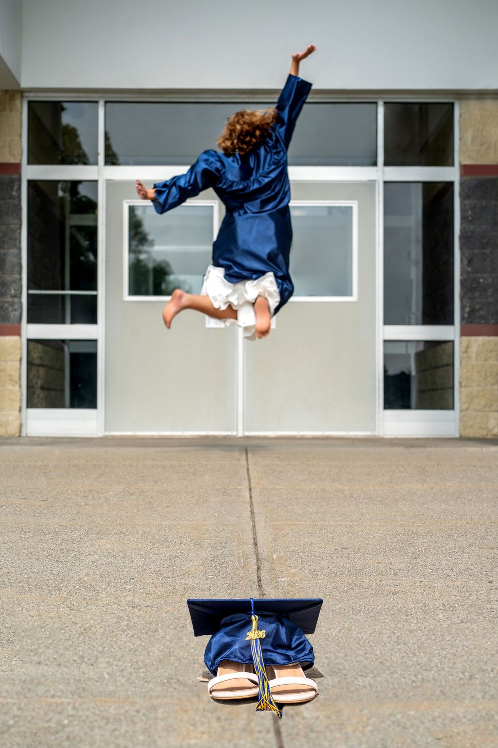 Child in blue graduation gown jumping joyfully outside school, cap and sandals on ground below.