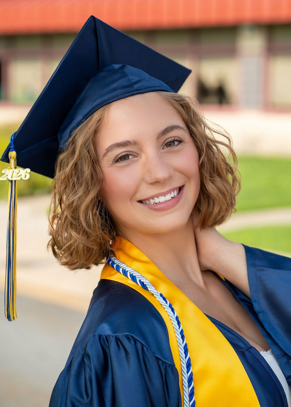 Smiling graduate in navy blue cap and gown with gold tassel and honor cords poses outdoors for senior portrait.