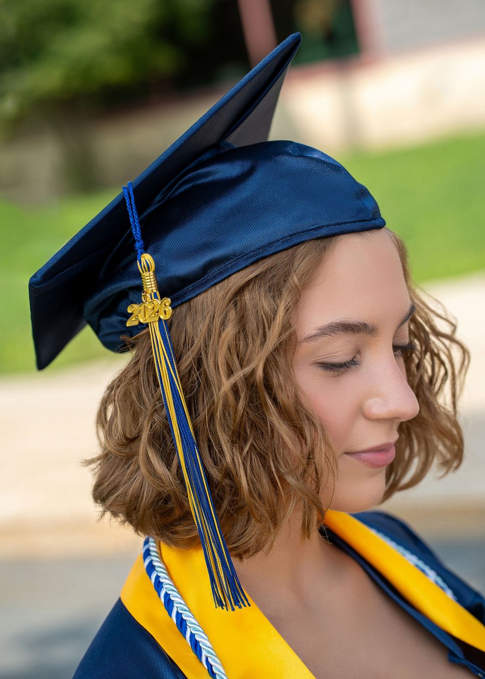 Young woman in navy blue graduation cap and gown with gold tassel and honor cords, posing outdoors.