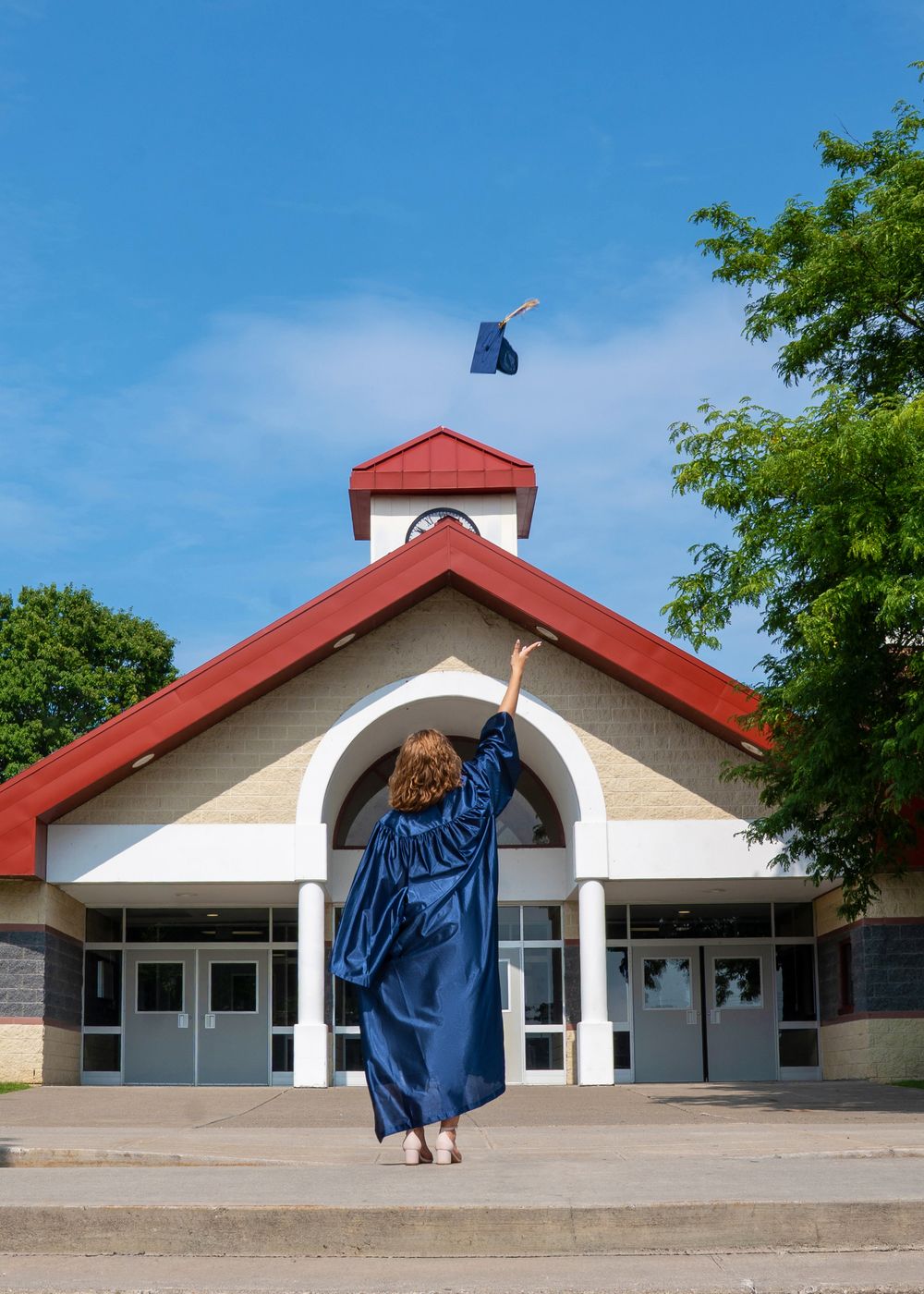 Graduate in blue gown tosses cap in front of school building under clear blue sky.