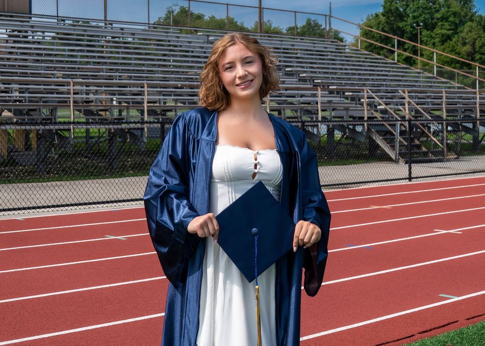 Young woman in blue graduation gown and cap holding diploma on red track field with bleachers behind her.