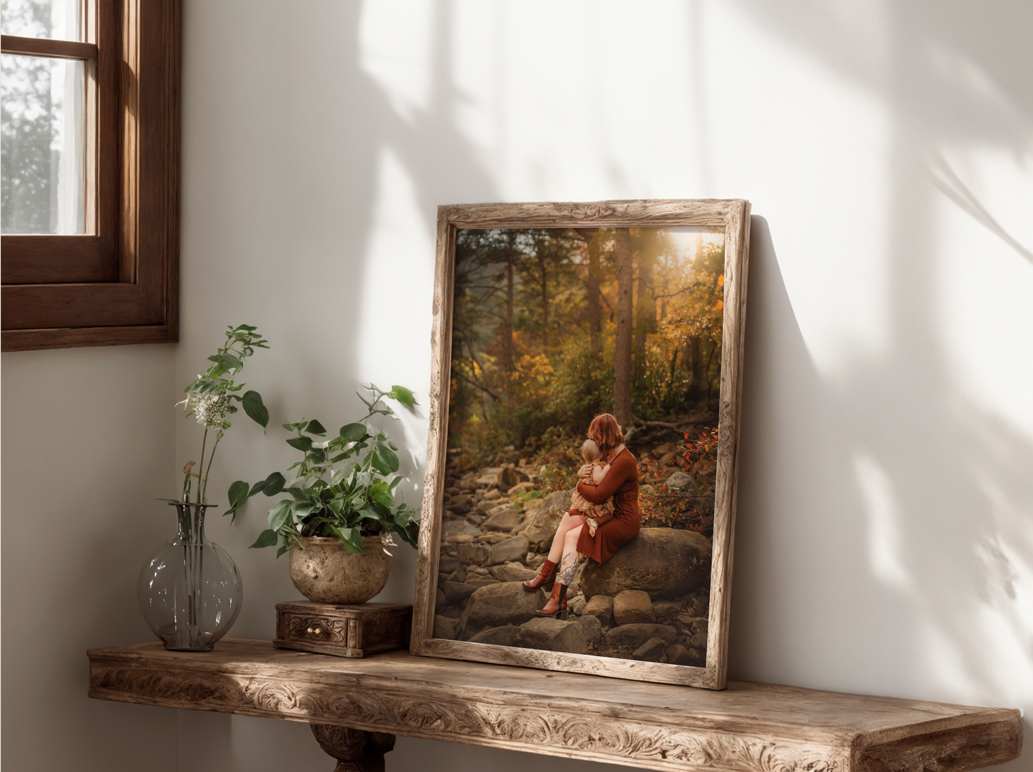 Framed autumn forest painting of a woman sitting on rocks, displayed on a rustic wooden shelf with green plants.