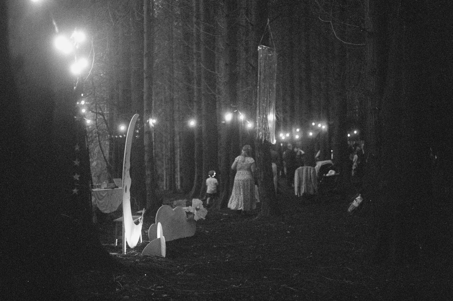 Black and white photo of an enchanting nighttime forest gathering with string lights and elegantly dressed guests.