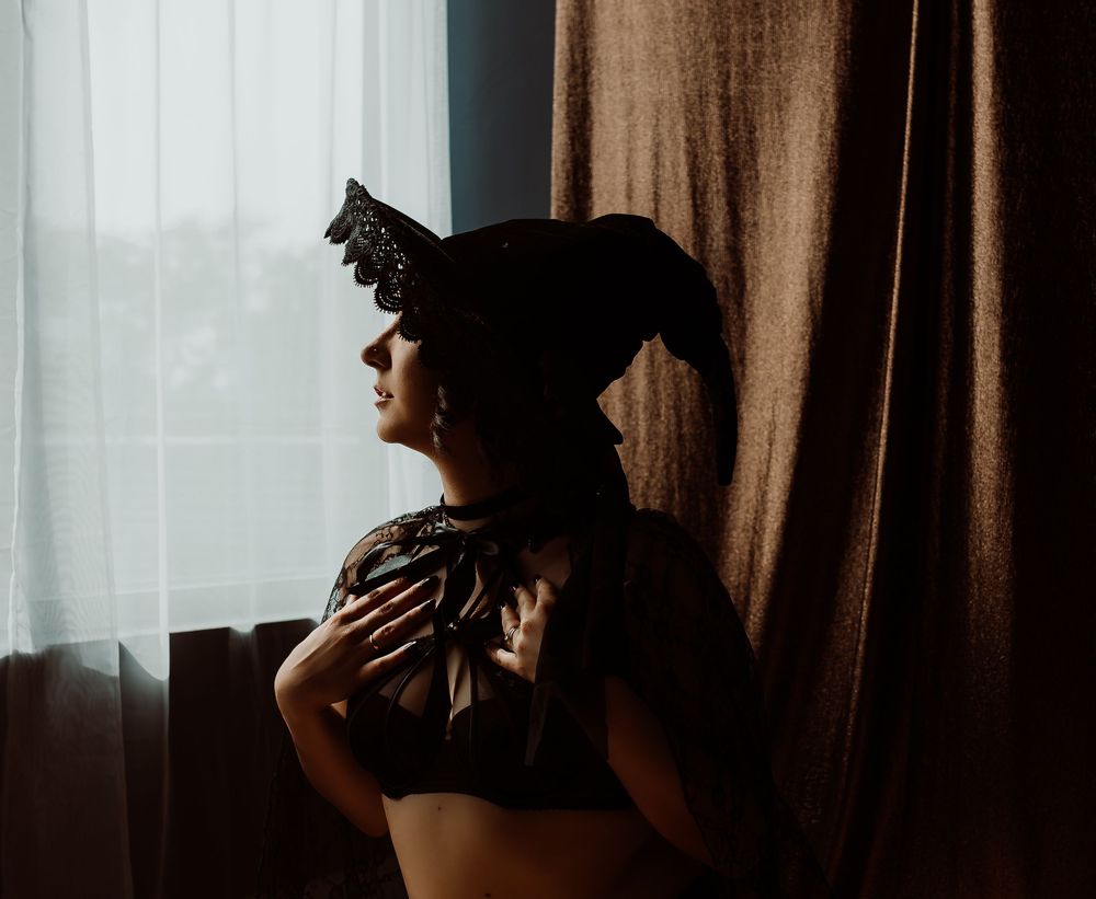 A woman doing a boudoir photoshoot in a dark lace bra and ornate black witch hat near a curtained window.