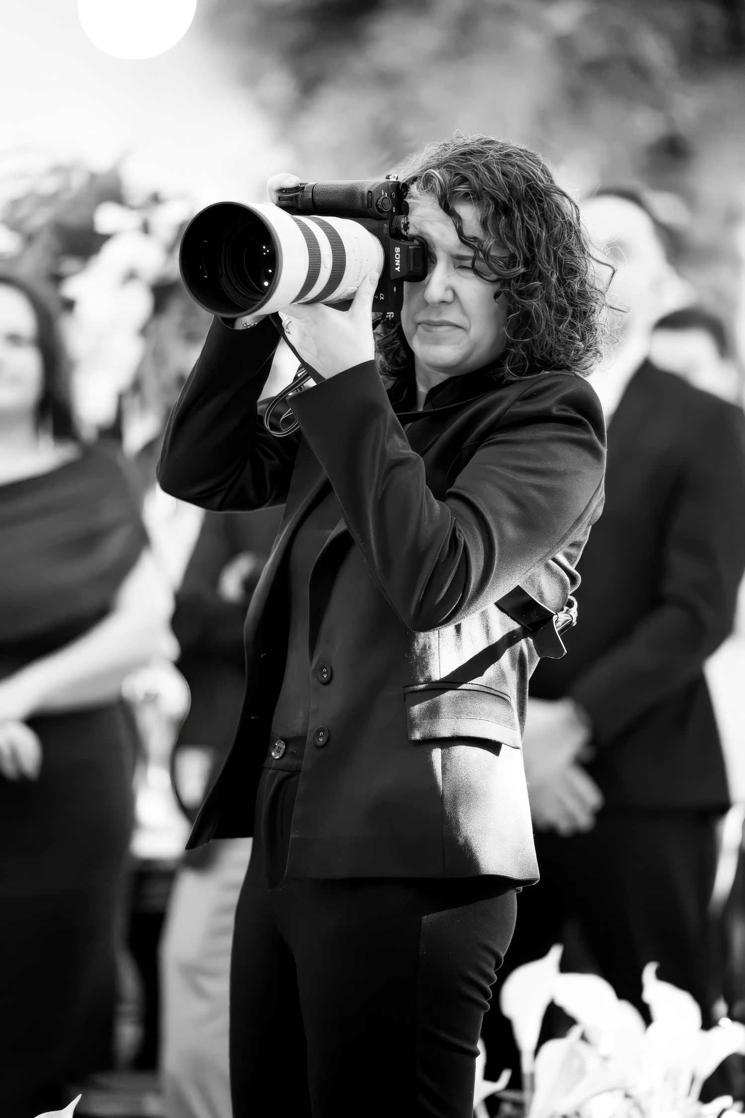 Black and white photo of a photographer with curly hair using a Sony camera with a large telephoto lens outdoors.
