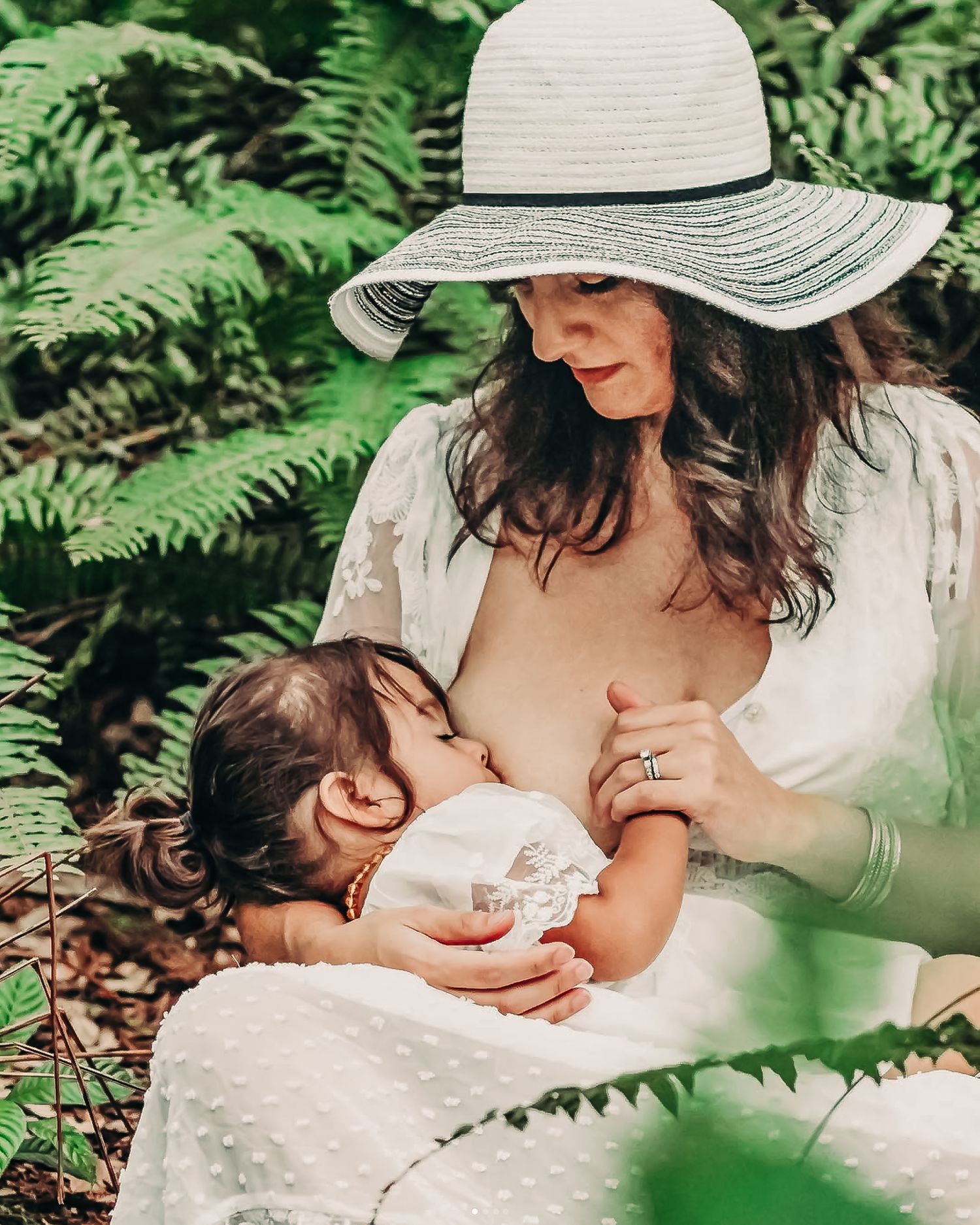 Mother in white dress and sun hat breastfeeding toddler outdoors surrounded by lush green ferns at Delray Oaks Natural Area.