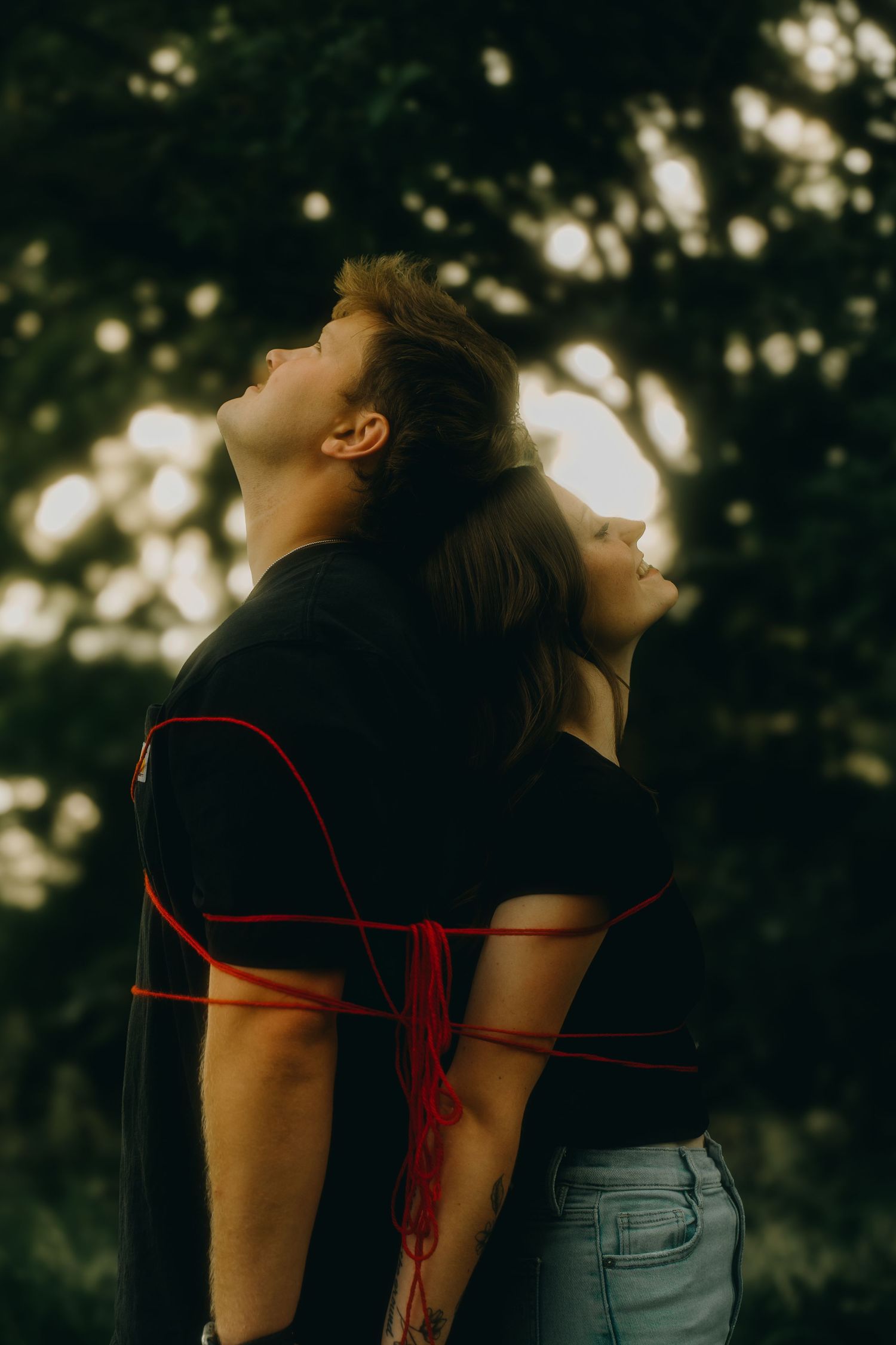 Couple bound together with red string standing back to back outdoors with bokeh tree background at golden hour.