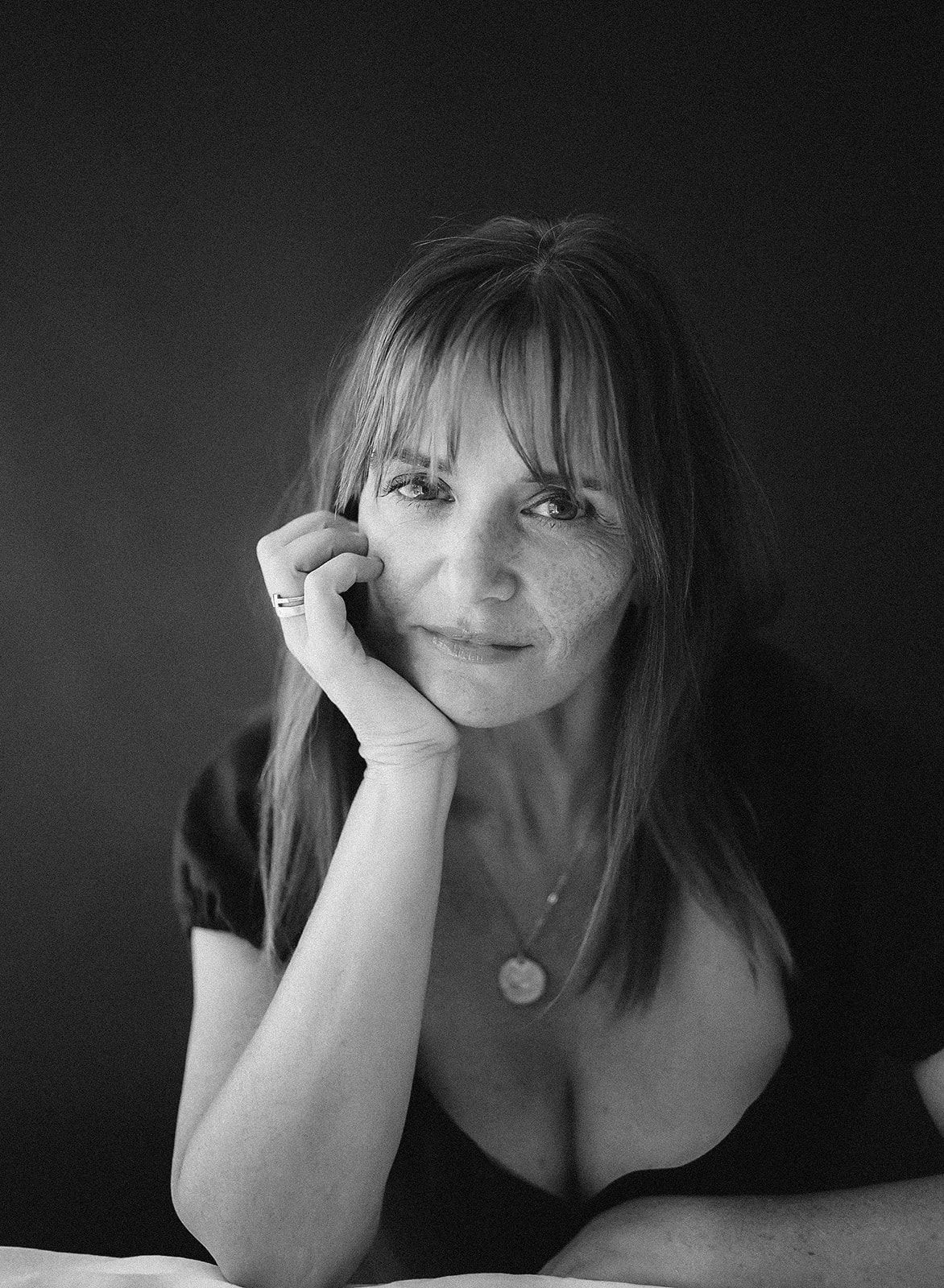 Black and white portrait of a woman with bangs resting her chin on her hand, wearing a necklace.