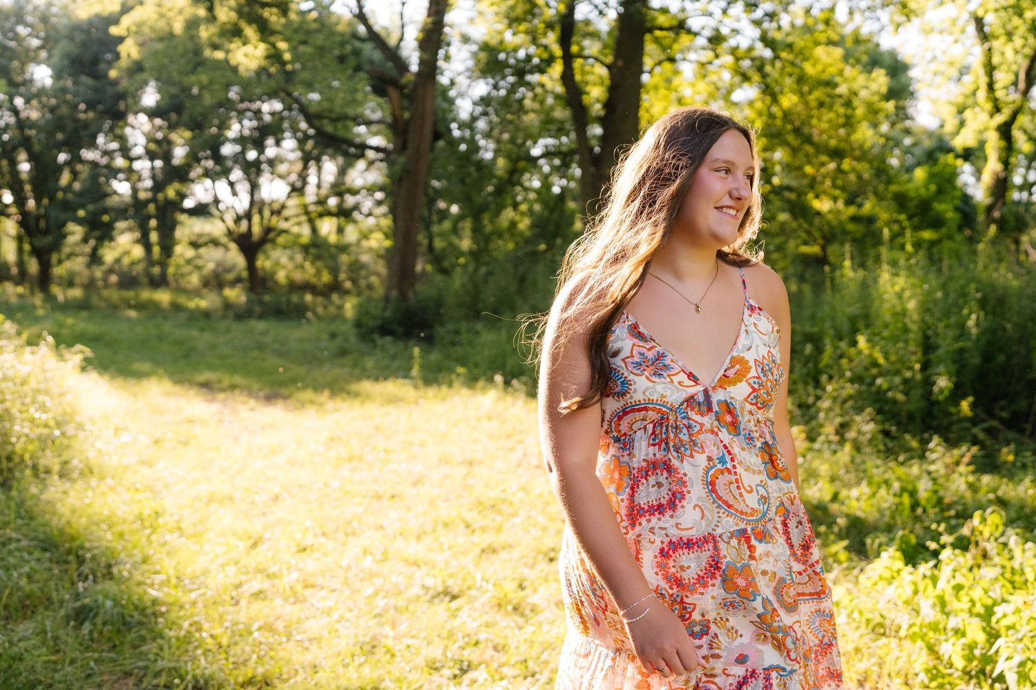 Young woman in floral sundress smiling outdoors in sunlit green field with trees in background.