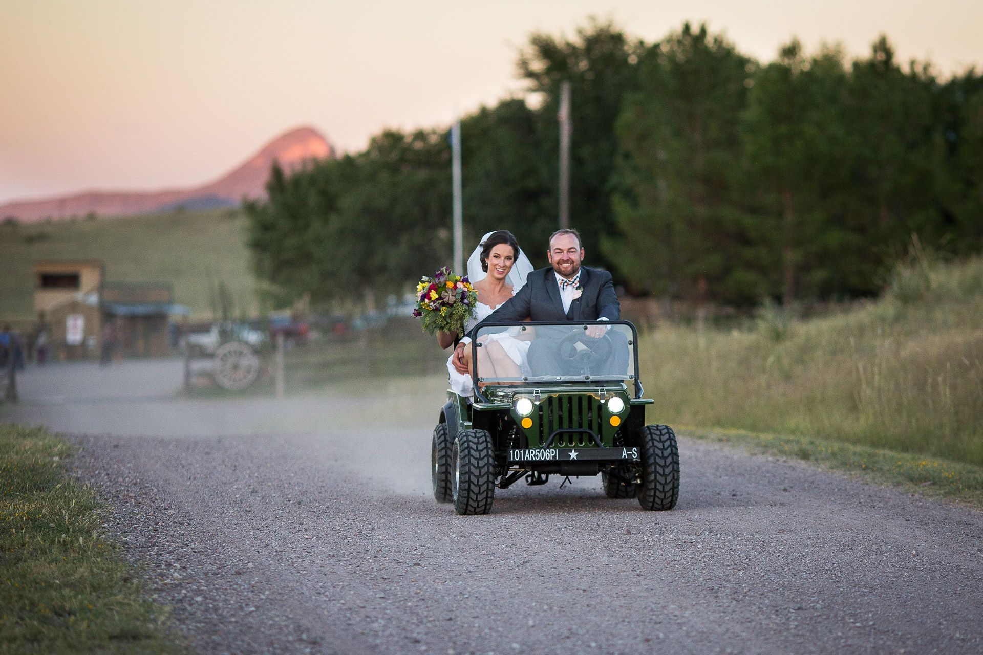 Bride and groom riding a vintage green Jeep down a gravel road at sunset during their outdoor wedding celebration.