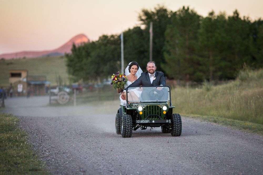 Bride and groom riding a vintage green Jeep down a gravel road at sunset during their outdoor wedding celebration.