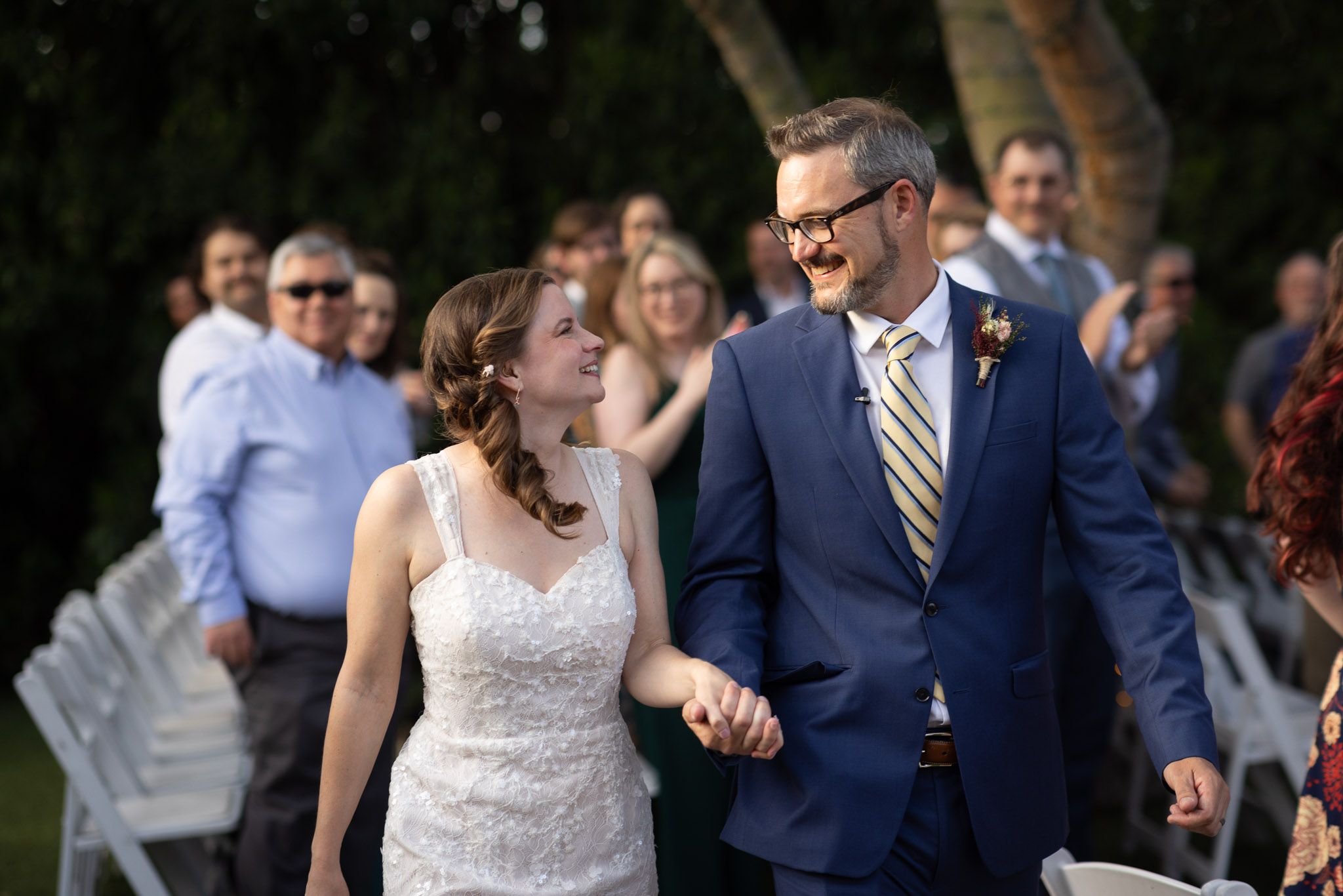 Newlywed couple walks down the aisle smiling, bride in white gown, groom in blue suit, outdoor wedding ceremony.