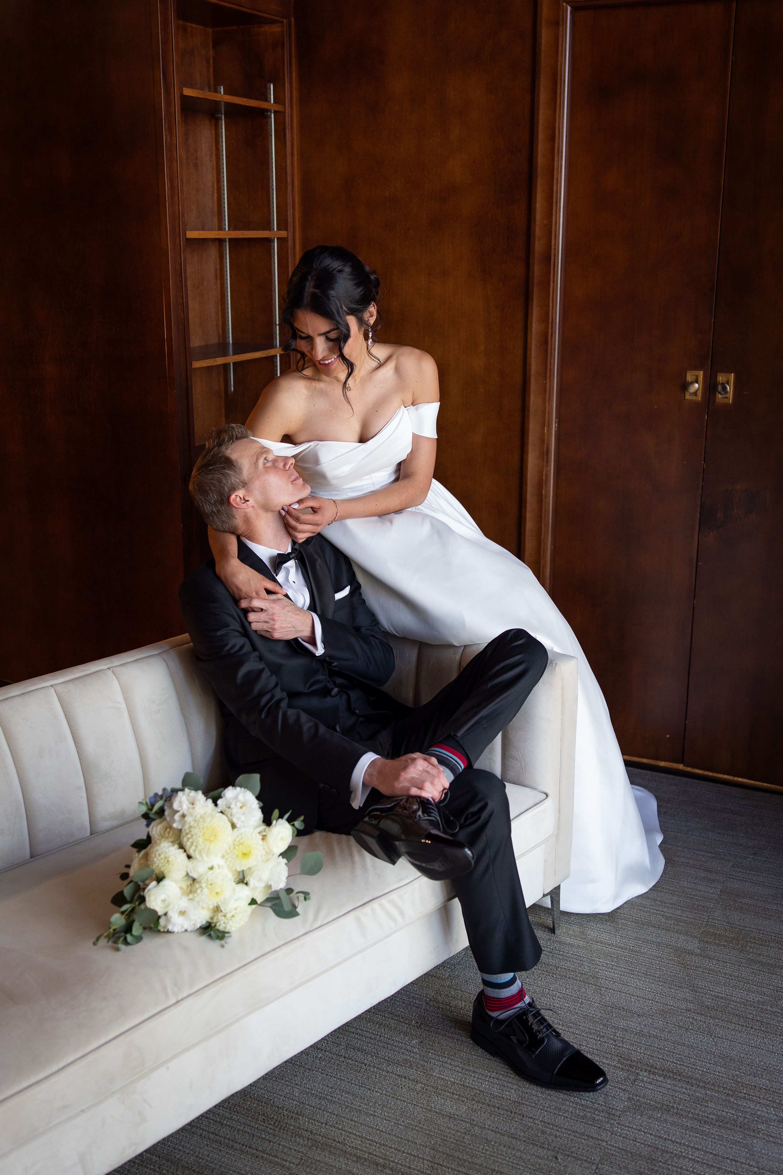 Bride in white off-shoulder gown sits with groom in black tuxedo on cream sofa, white rose bouquet nearby.
