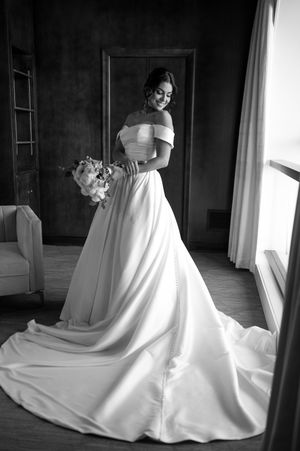 Smiling bride in off-shoulder white gown with long train holds bouquet in black and white photo.