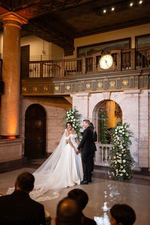 Bride and groom hold hands at elegant indoor wedding ceremony with floral arch and ornate balcony backdrop.