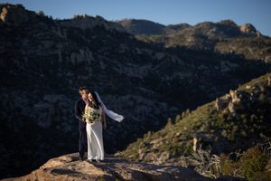 Bride and groom embrace on rocky mountain summit with stunning desert peaks backdrop during outdoor wedding ceremony.
