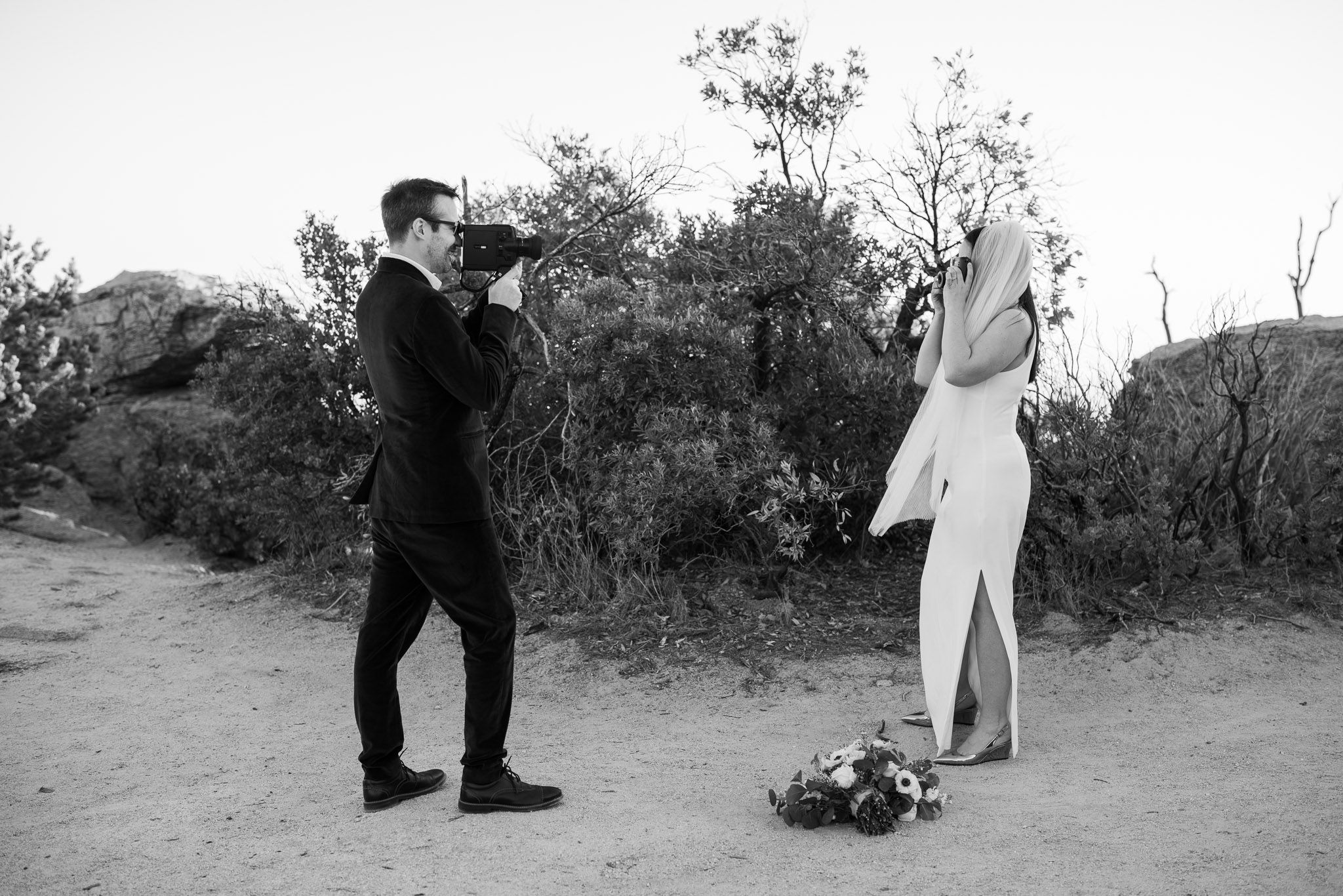 Black and white photo of a groom photographing his bride outdoors on a dirt path surrounded by trees.