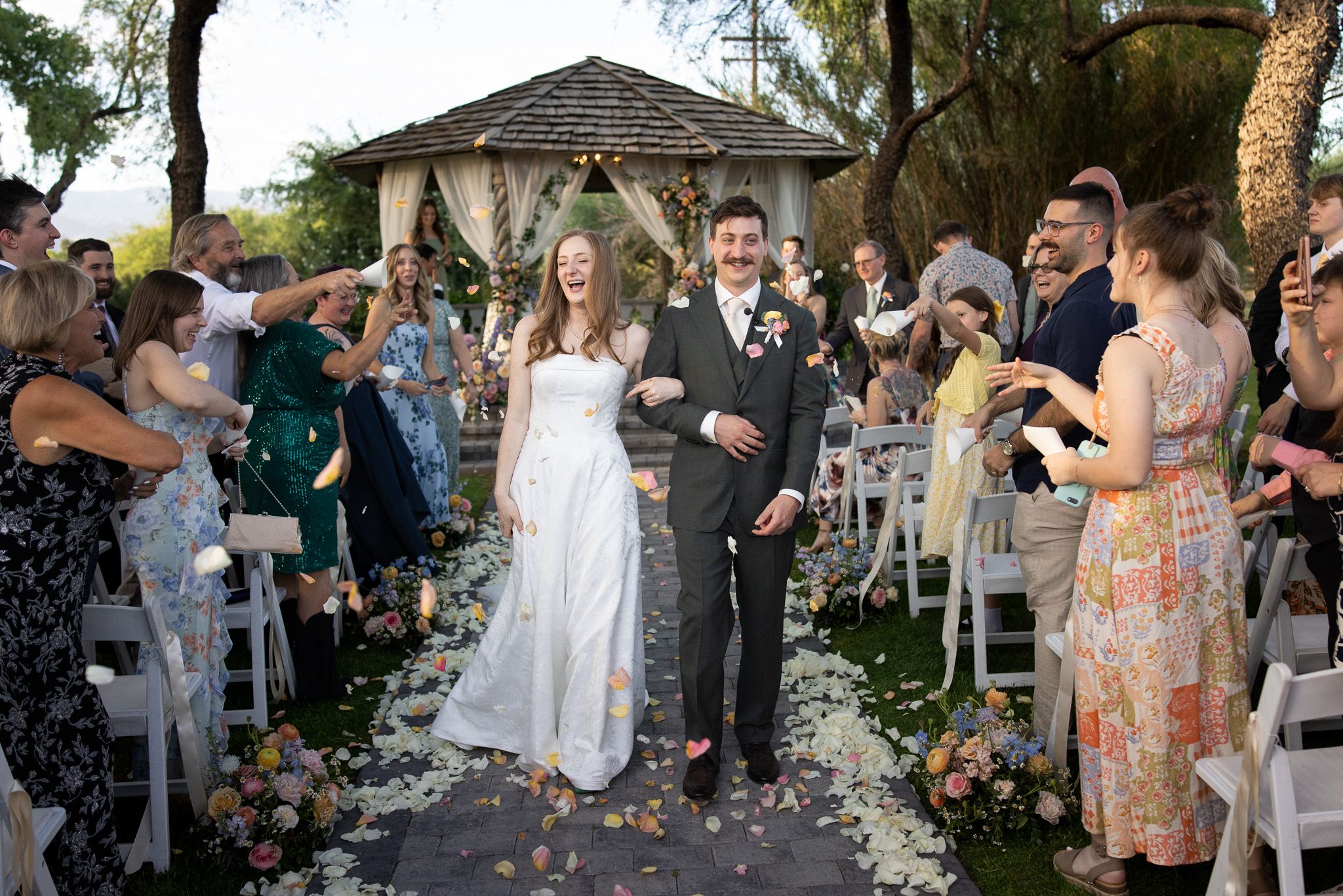 Bride and groom walk down the aisle as guests toss flower petals at an outdoor garden wedding ceremony.