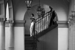 Black and white photo of two elegantly dressed people on a grand staircase with arched columns and a hanging lantern.