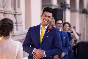Smiling groom in navy suit and yellow tie reacts emotionally during outdoor wedding ceremony with bridal party in background.