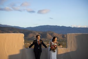Bride and groom hold hands on a rooftop with stunning mountain views and golden sunset sky in New Mexico.