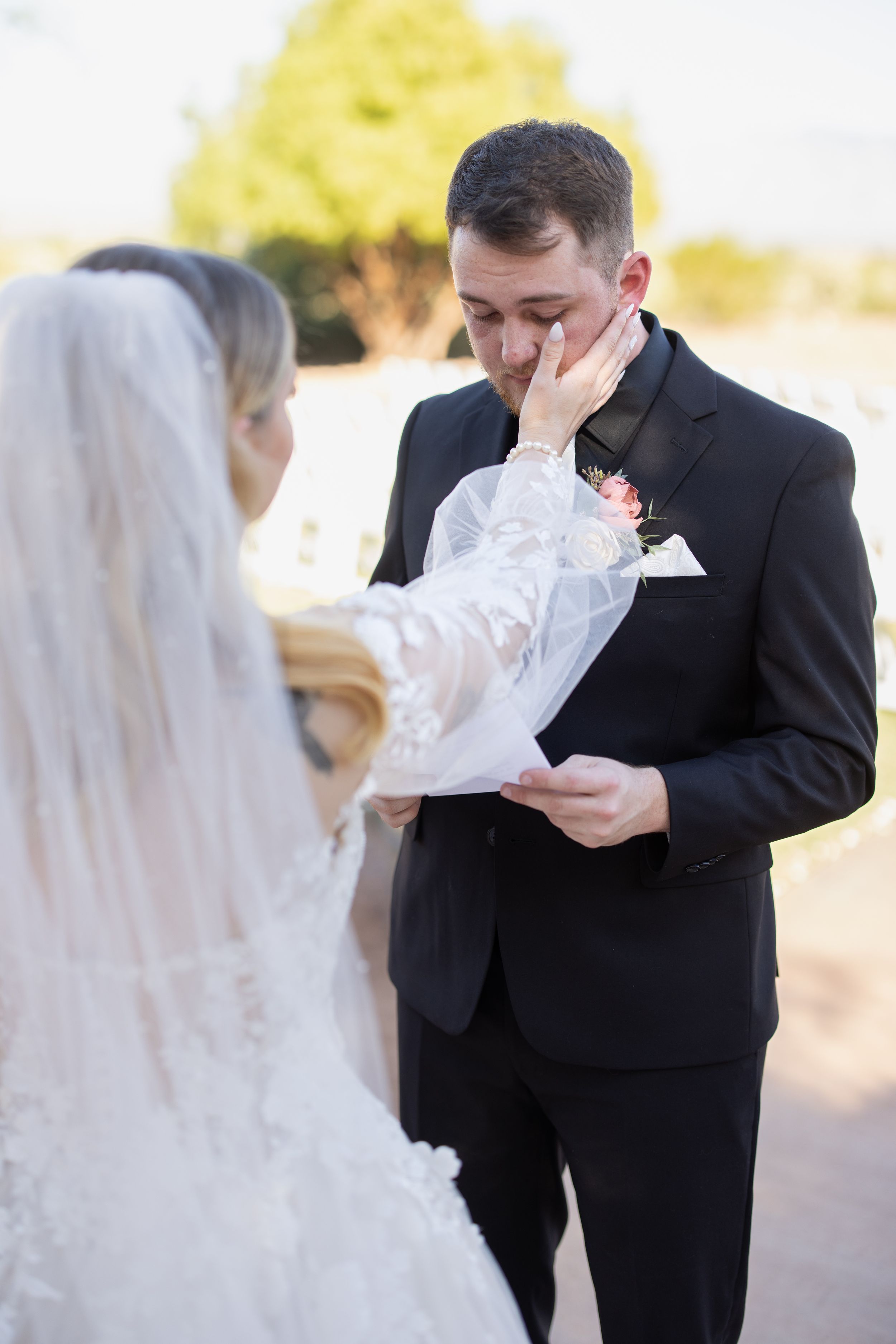 Emotional groom wiping tears as bride touches his face during outdoor wedding ceremony vow exchange.