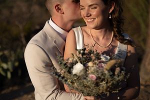 A groom in a beige suit kisses his smiling bride holding a rustic bouquet of wildflowers outdoors.