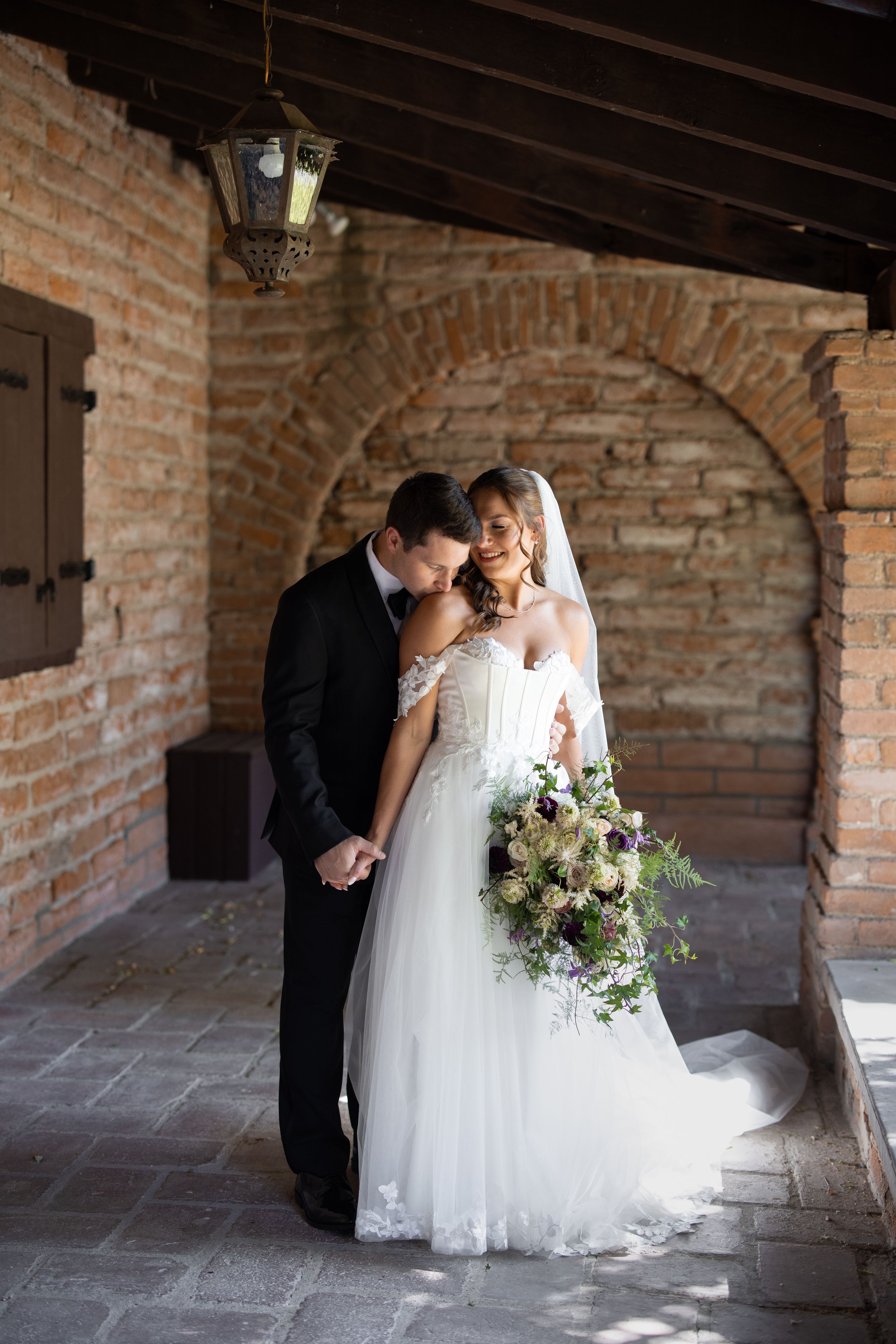 Bride and groom embrace under rustic brick archway, bride holding elegant bouquet in white gown and veil.