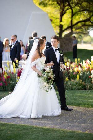 Bride in white gown and groom in tuxedo holding champagne glasses at outdoor wedding with colorful garden backdrop.