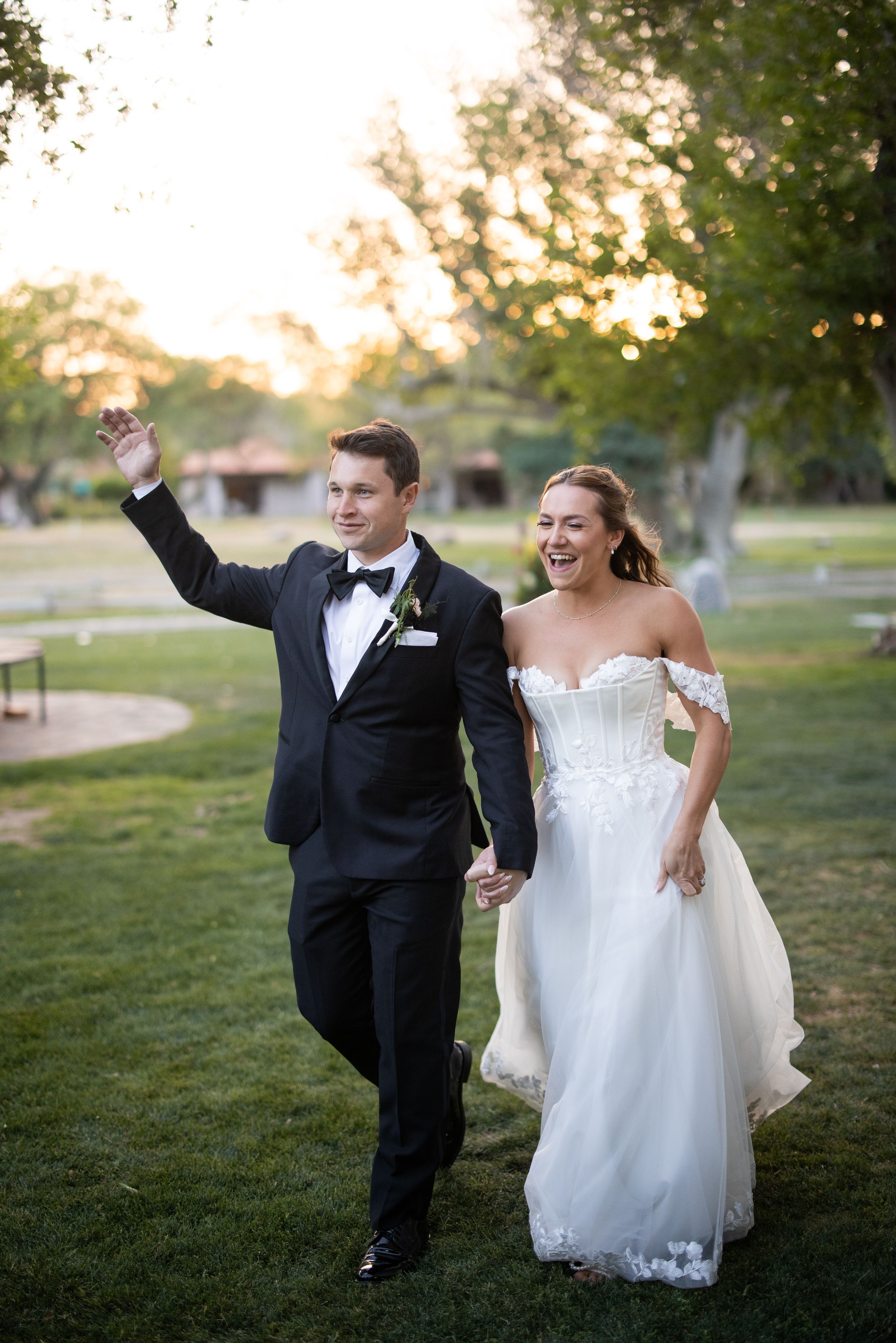 Joyful bride and groom walking hand in hand outdoors at sunset, groom in tuxedo waving, bride in off-shoulder gown.
