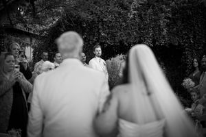 Black and white wedding photo of bride and groom walking toward smiling officiant outdoors.