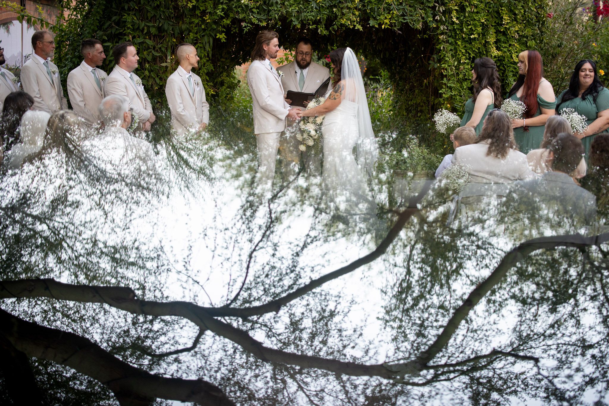 Wedding ceremony outdoors with couple exchanging vows, bridal party in white, framed by lush green ivy and tree branches.